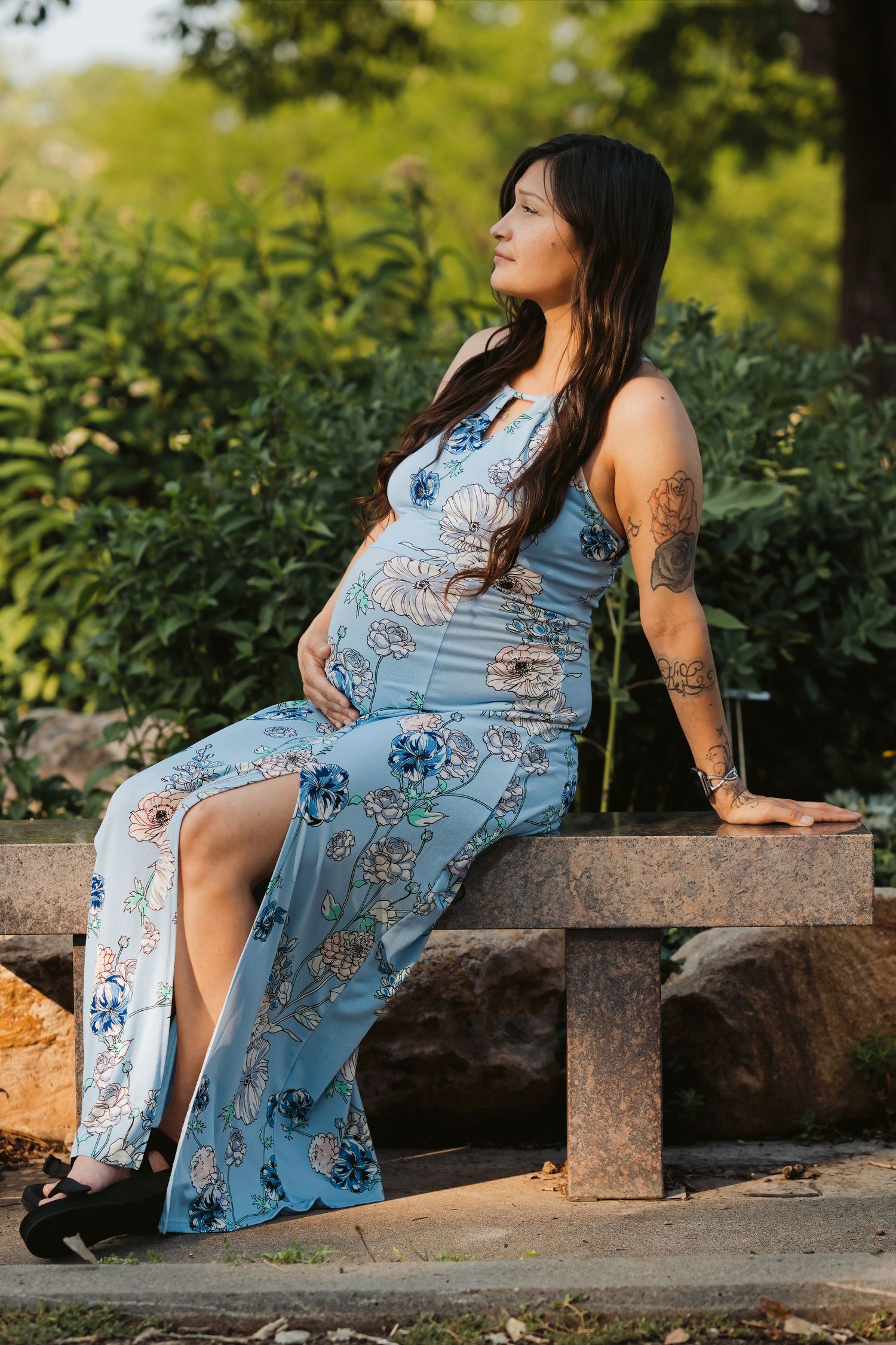 Pregnant woman in blue floral maxi dress sitting on stone bench in garden, showing baby bump with tattoos visible.