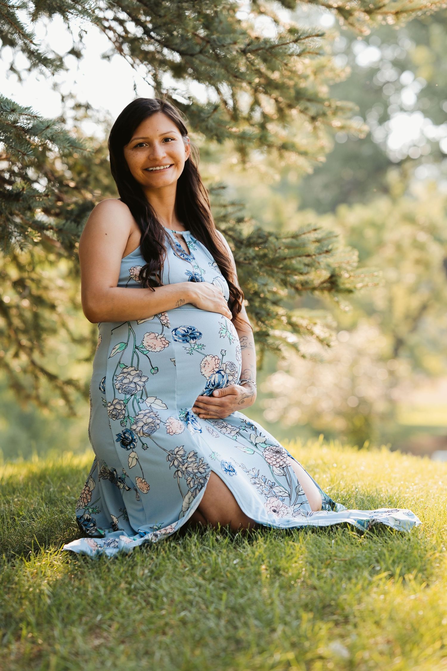 Smiling pregnant woman in floral blue dress sitting on grass, cradling baby bump outdoors near evergreen tree.