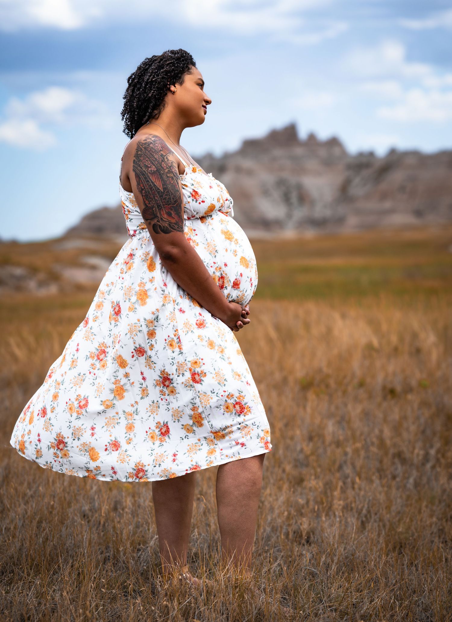 Pregnant woman in floral dress standing in a grassy field with rocky badlands formations in the background.