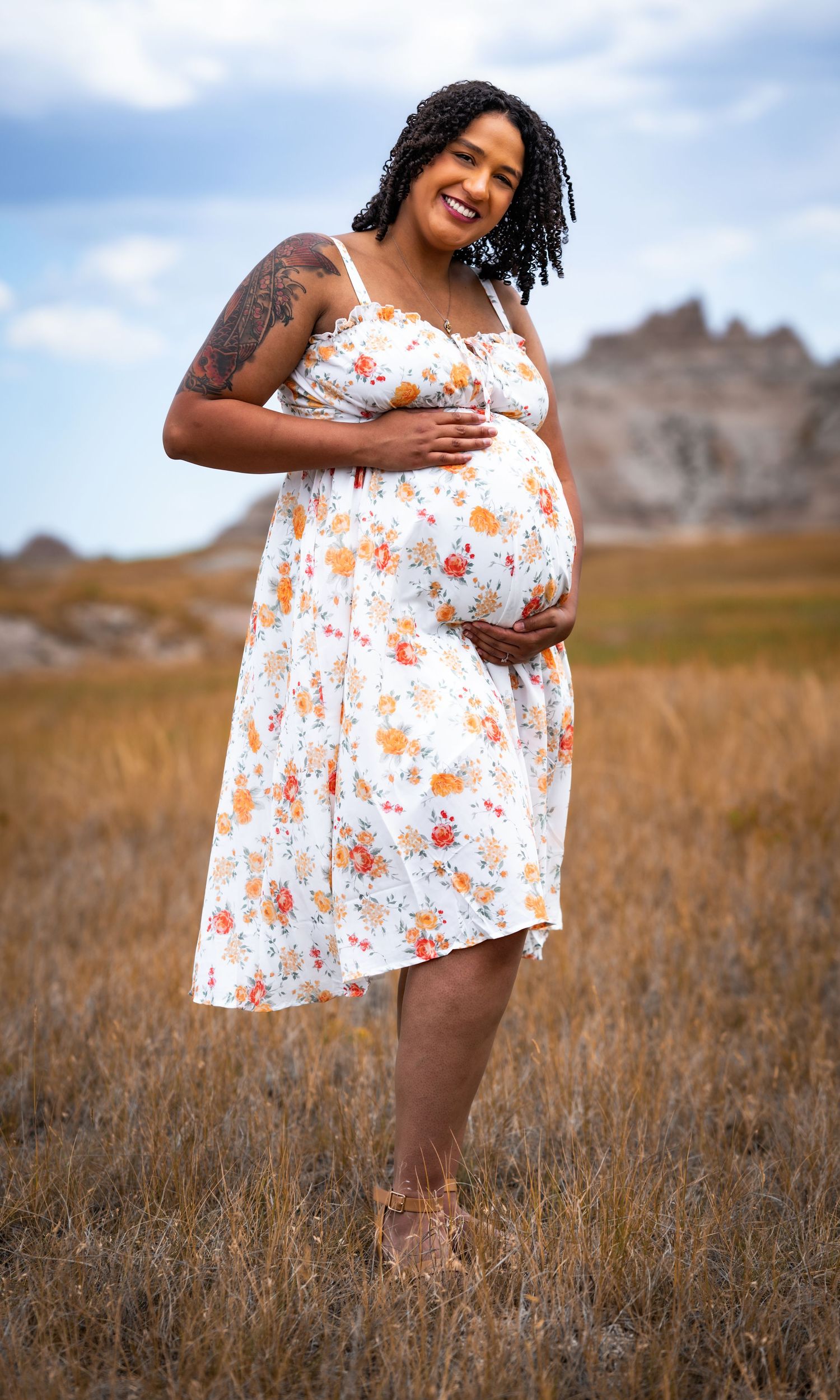 Smiling pregnant woman in floral dress holding belly in golden field with rocky formations backdrop.