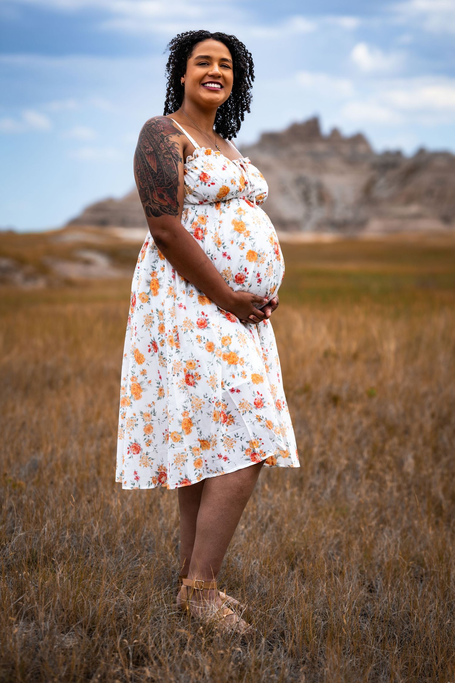 Smiling pregnant woman in floral dress standing in grassy field with rocky badlands formation in background.