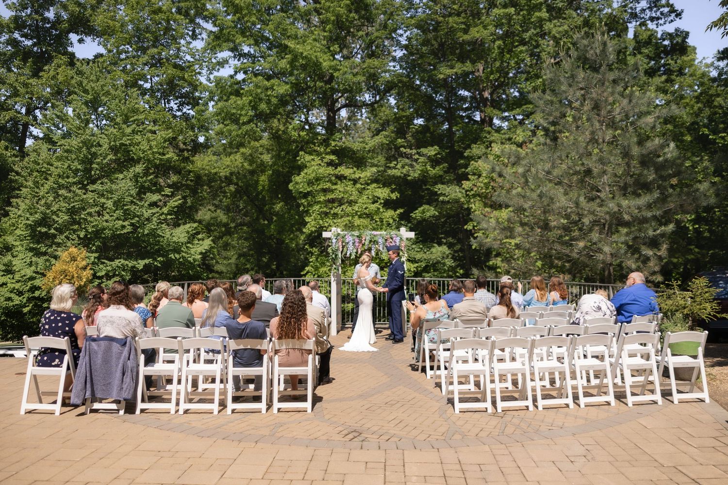 Outdoor wedding ceremony with guests seated in white chairs on a brick patio surrounded by lush green trees.