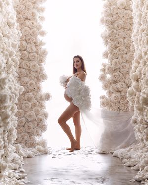 Modern maternity photo: mum wearing a unique white rose cape-style gown in studio, backlit with white rose walls.