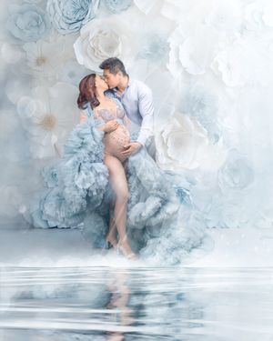 A whimsical maternity portrait featuring a man leaning over and kissing his pregnant wife against a blue and white flower wall and water.