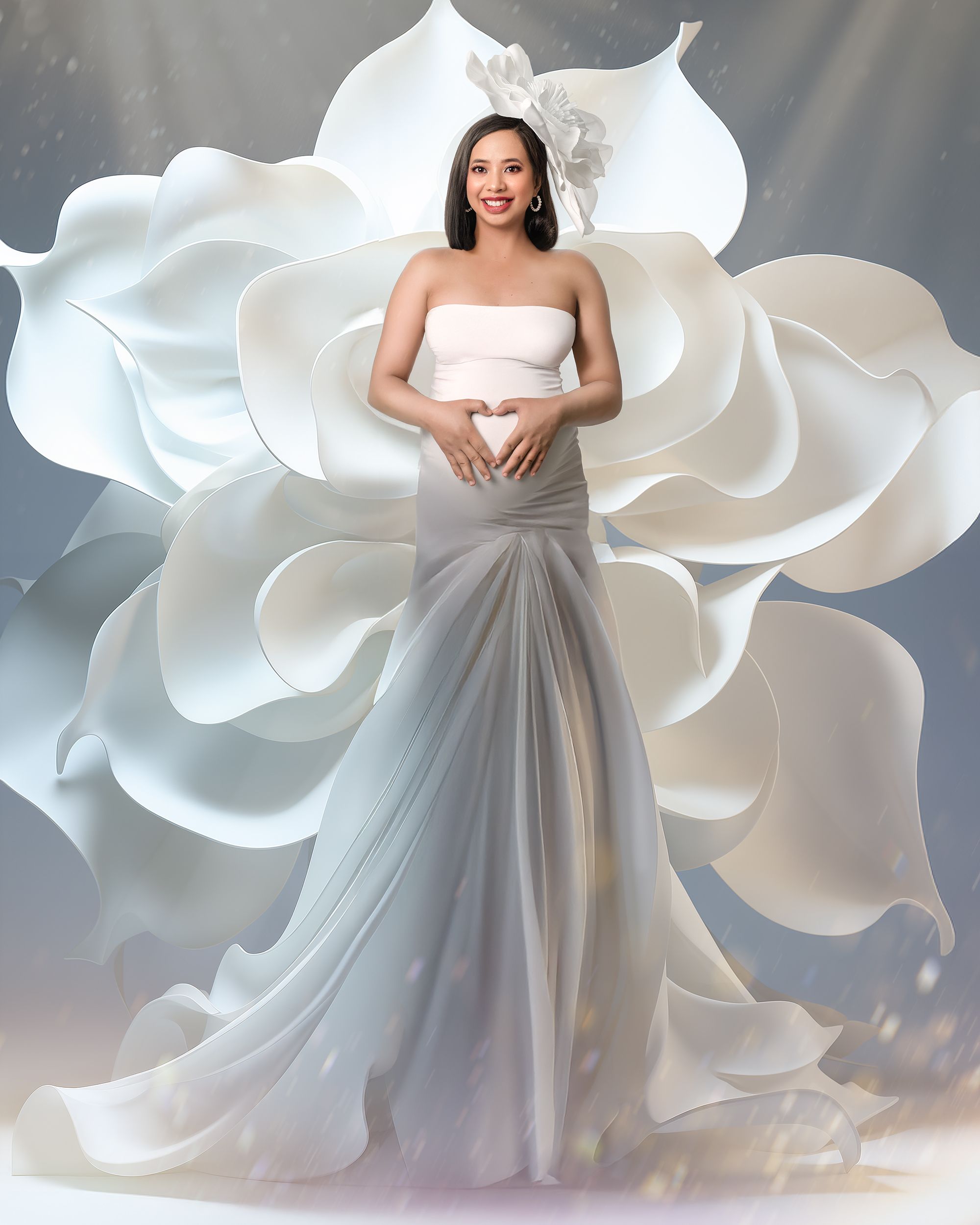 A pregnant woman poses making a heart shape with her hands over her belly, glowing looking ethereal in long white gown against giant flower.