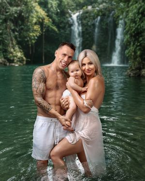 A Mother and Father hold their baby daughter lovingly all wearing white, dripping wet standing in a lush green waterfall scene.