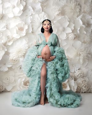 A mum to be smiles warmly wearing sage green couture robe against a white flower wall looking beautiful.