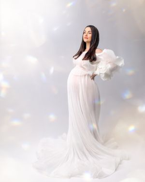 Woman wears white matching pleat modern styled gown against white backdrop light beams sparkle around her, she smiles and looks divine.