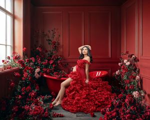 Exquisite pregnancy portrait of a woman in a large fluffy red designer gown sitting on a red bath surrounded by flowers.