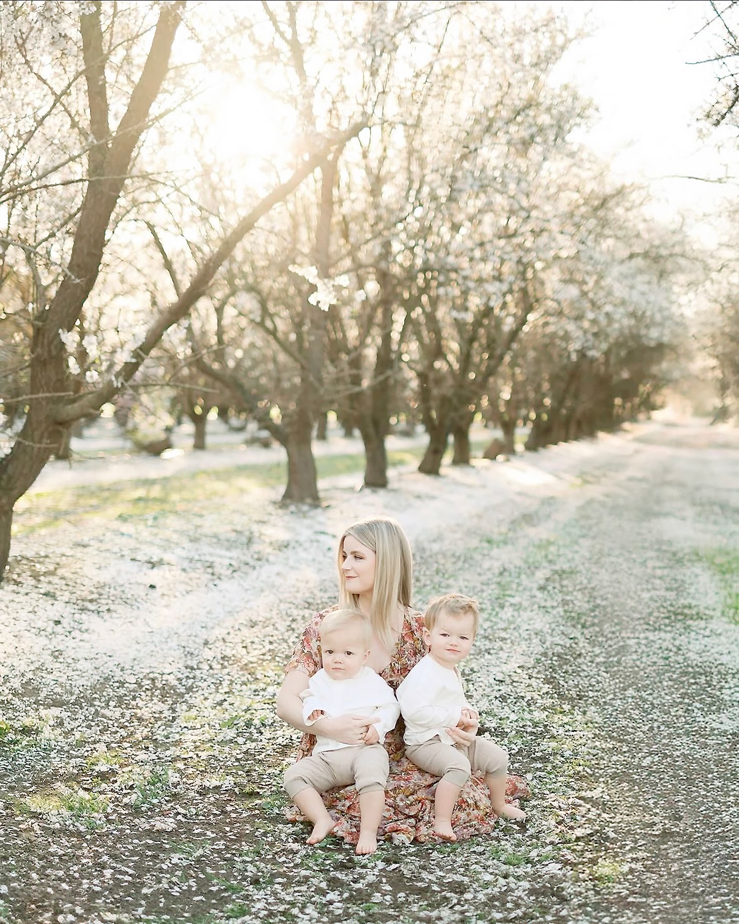 A mother sits with two toddlers in a sunlit orchard with white blossoms on the ground.