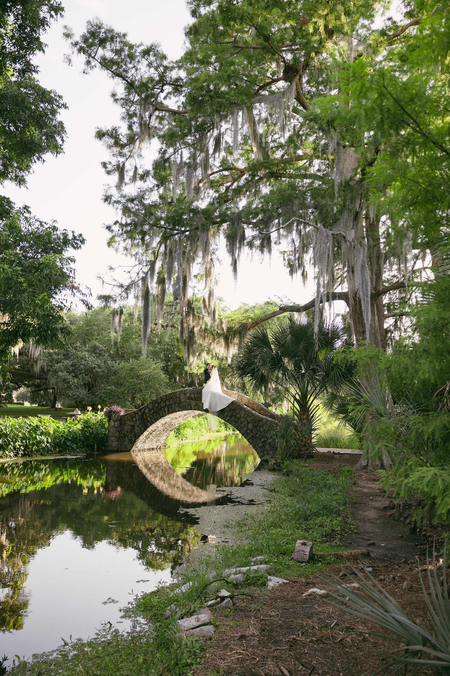 A bride in a white gown stands on a stone arch bridge surrounded by lush greenery and Spanish moss-draped trees.