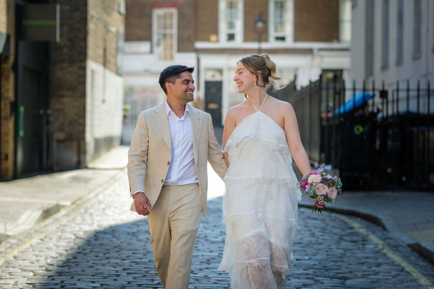 A bride and groom walk hand-in-hand on a cobblestone street, she in a white gown holding a bouquet, he in a tan suit.