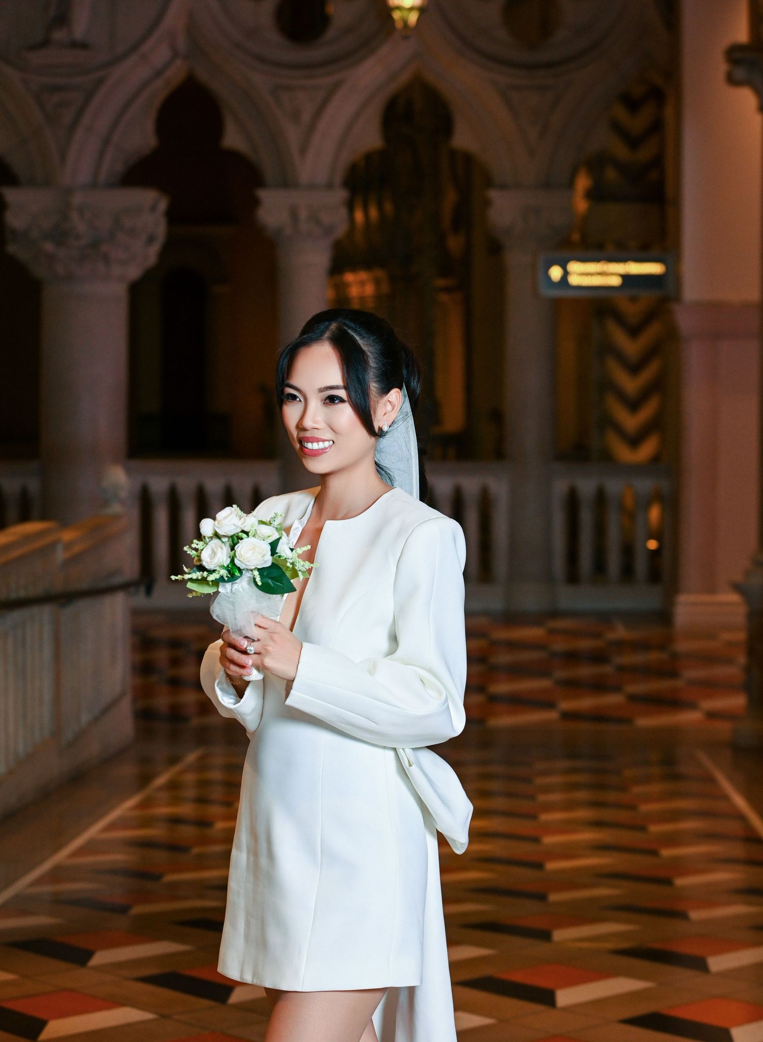 A smiling bride in a white mini dress holds a bouquet of white roses in an elegant arched interior.