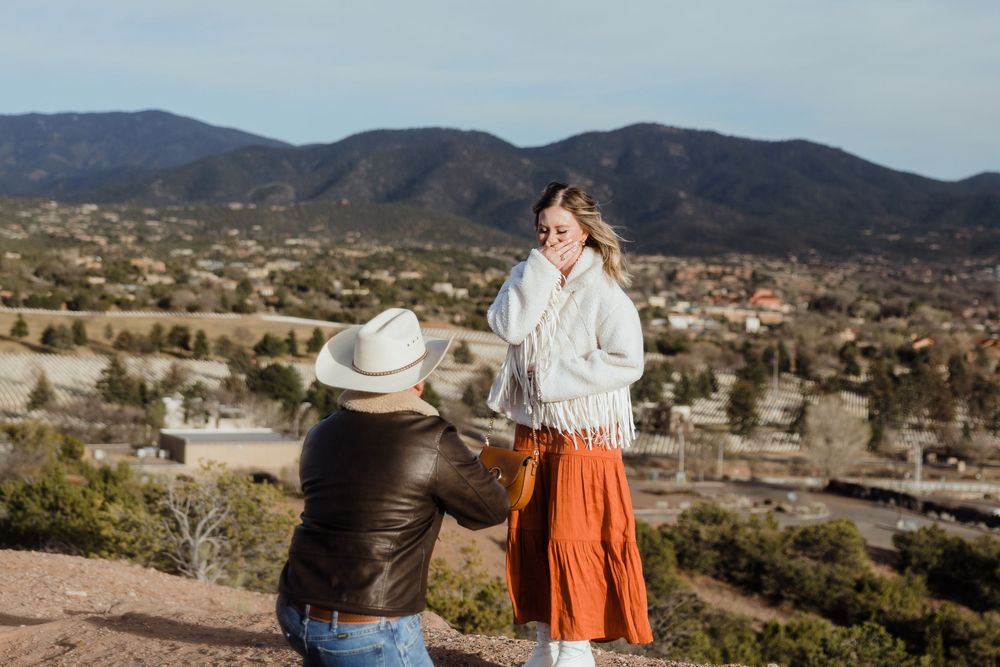 Man on one knee proposing to a woman in an orange skirt with mountain views behind them.