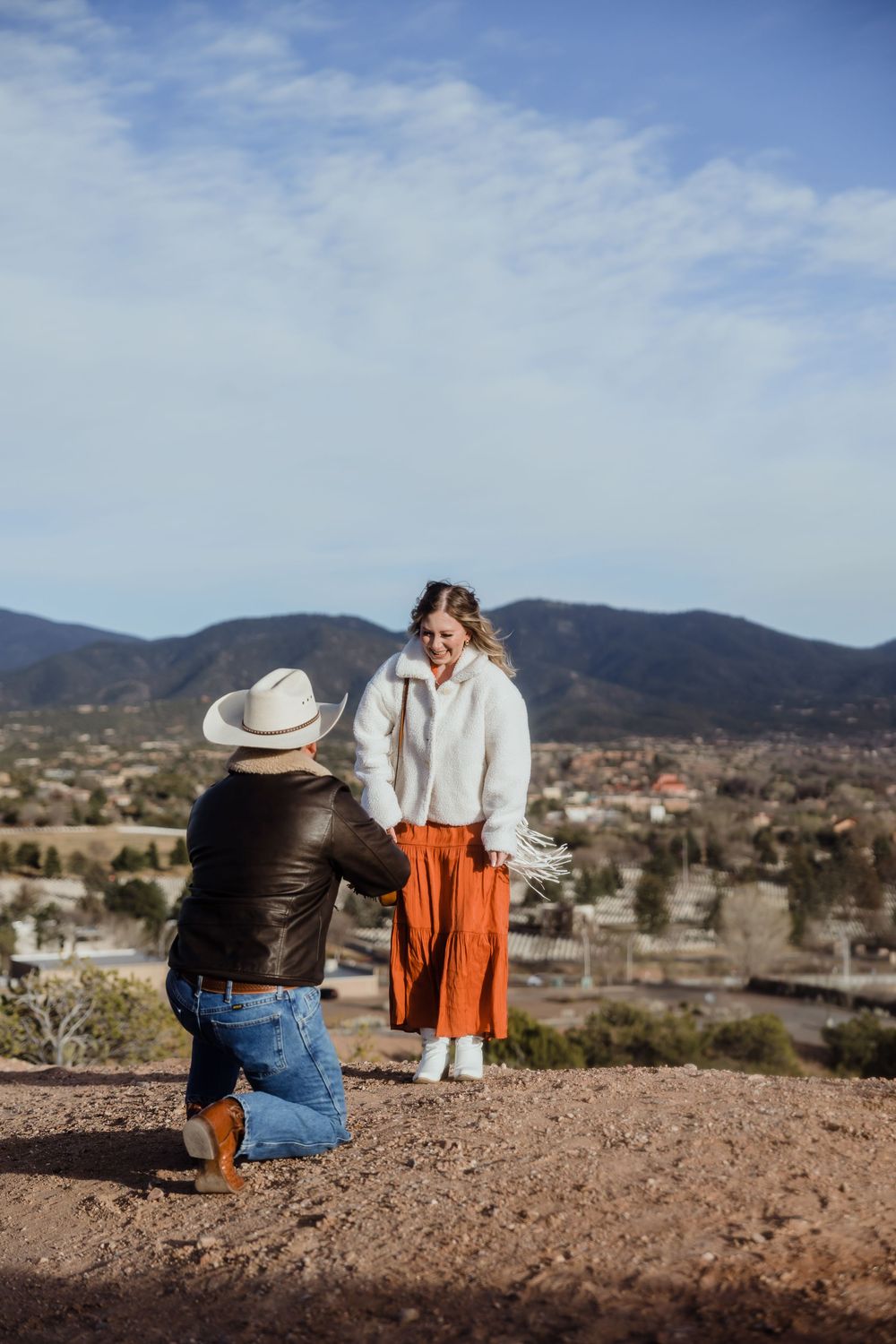 Man on one knee proposing to a woman on a hilltop overlooking a mountain town under a blue sky.