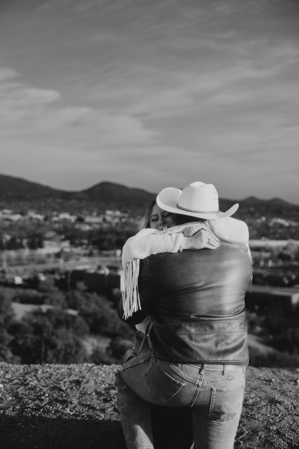 Black and white photo of couple embracing overlooking a scenic valley, woman in white fringe sweater and cowboy hat.