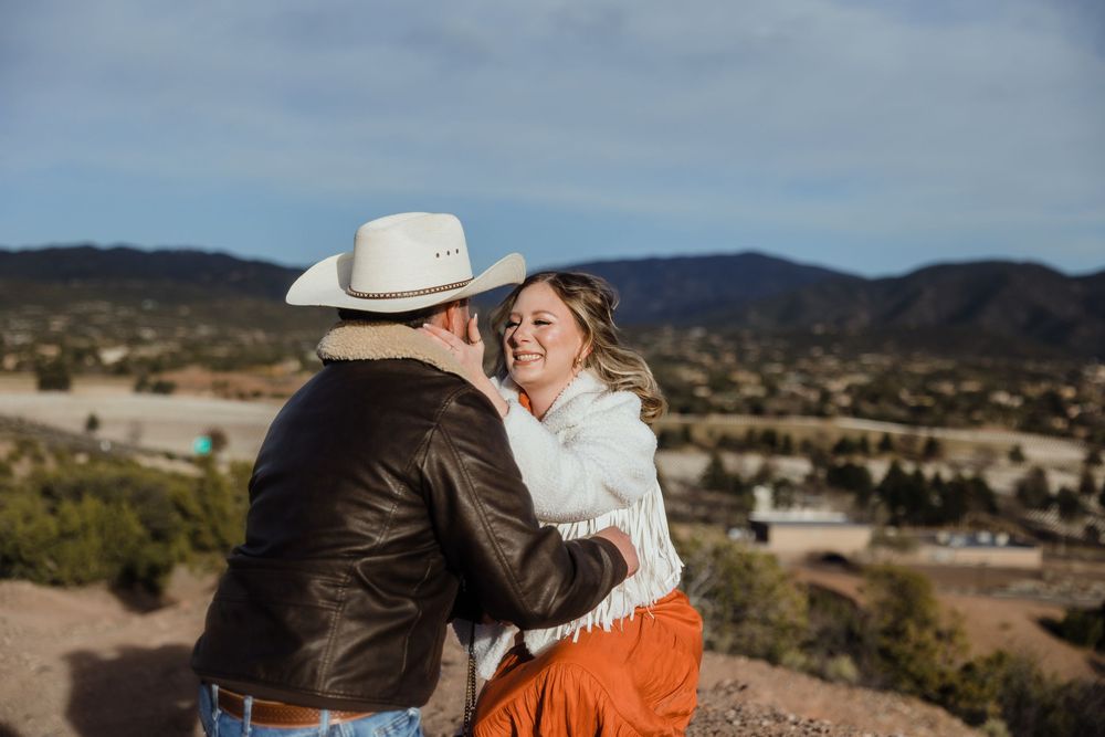 Couple laughing and embracing outdoors with mountain landscape, man in cowboy hat and brown leather jacket.