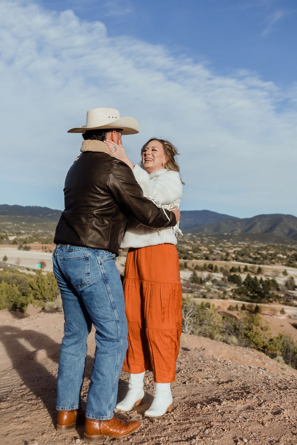 Happy couple embracing outdoors, man in cowboy hat and leather jacket, woman in orange pants laughing, mountain views behind.