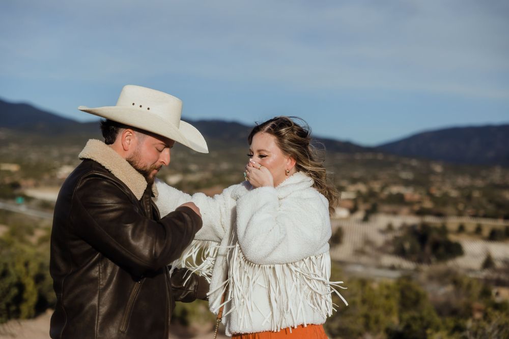 Man in white cowboy hat kissing woman's hand outdoors with mountain valley backdrop, both in western attire.