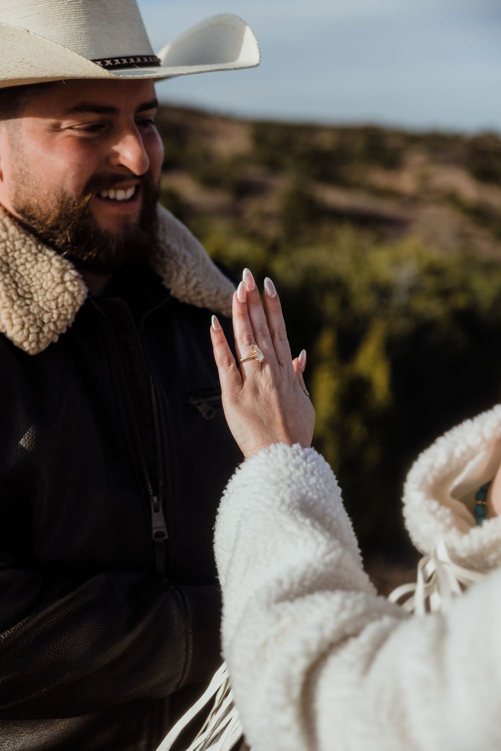 Bearded man in cowboy hat smiling at womans hand displaying a gold engagement ring outdoors.