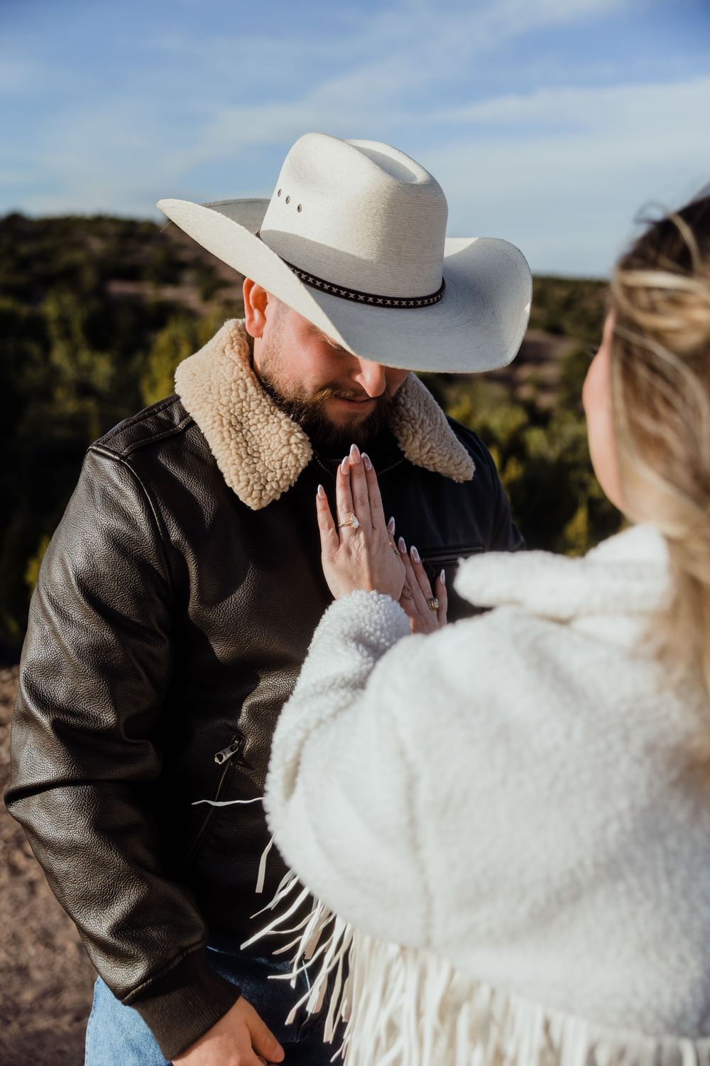 Man in cowboy hat kisses woman's hand with engagement ring outdoors in scenic landscape.