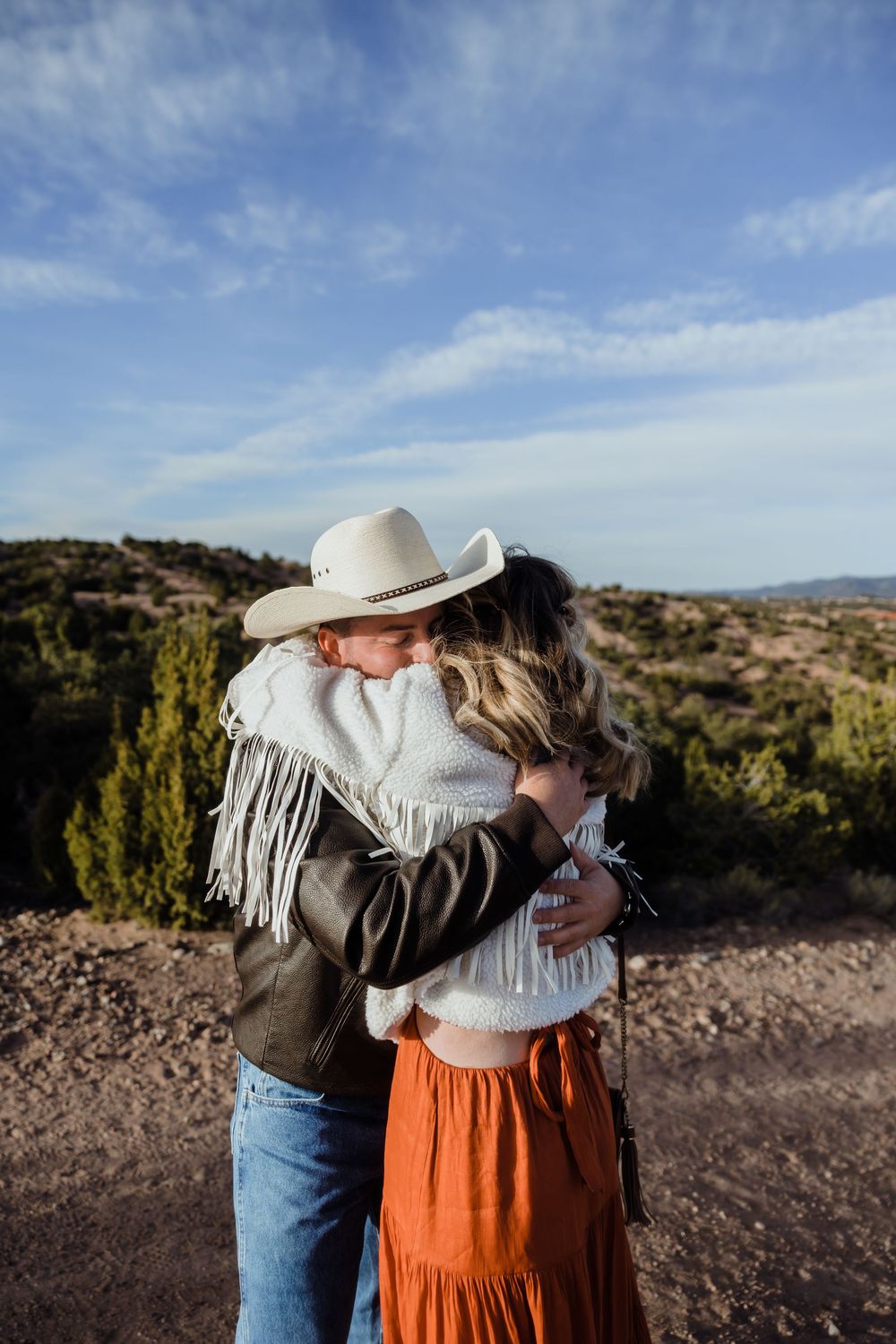 Couple sharing a romantic hug outdoors, woman in orange skirt and white fringe wrap, man in cowboy hat.