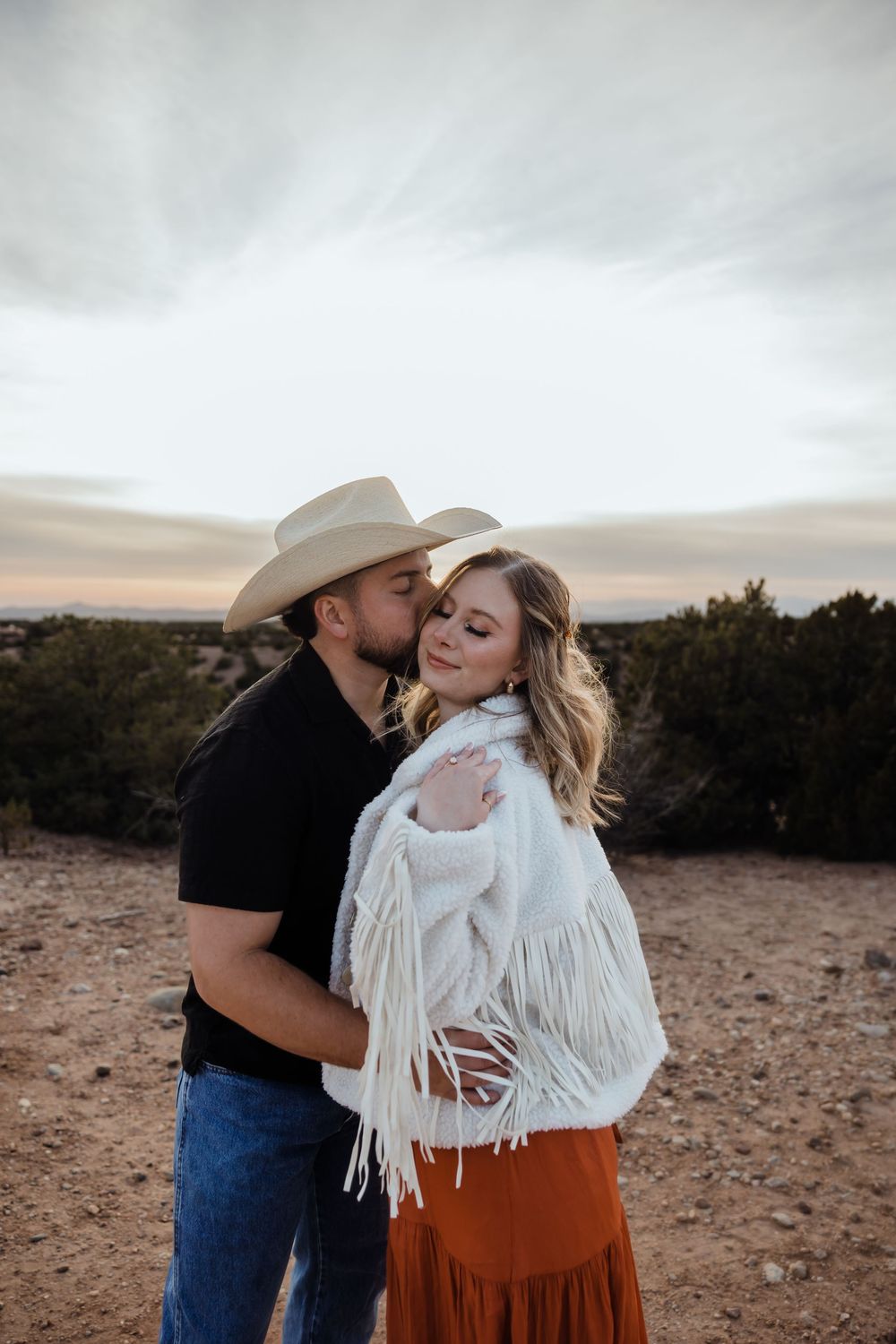 Man in cowboy hat kissing woman's cheek, she wears white fringe jacket and rust skirt, desert sunset backdrop.