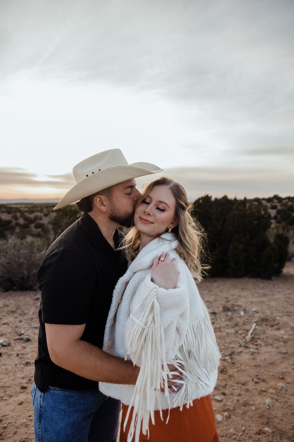 Couple embracing at sunset in desert, man in cowboy hat kissing woman in white fringe jacket and rust skirt.