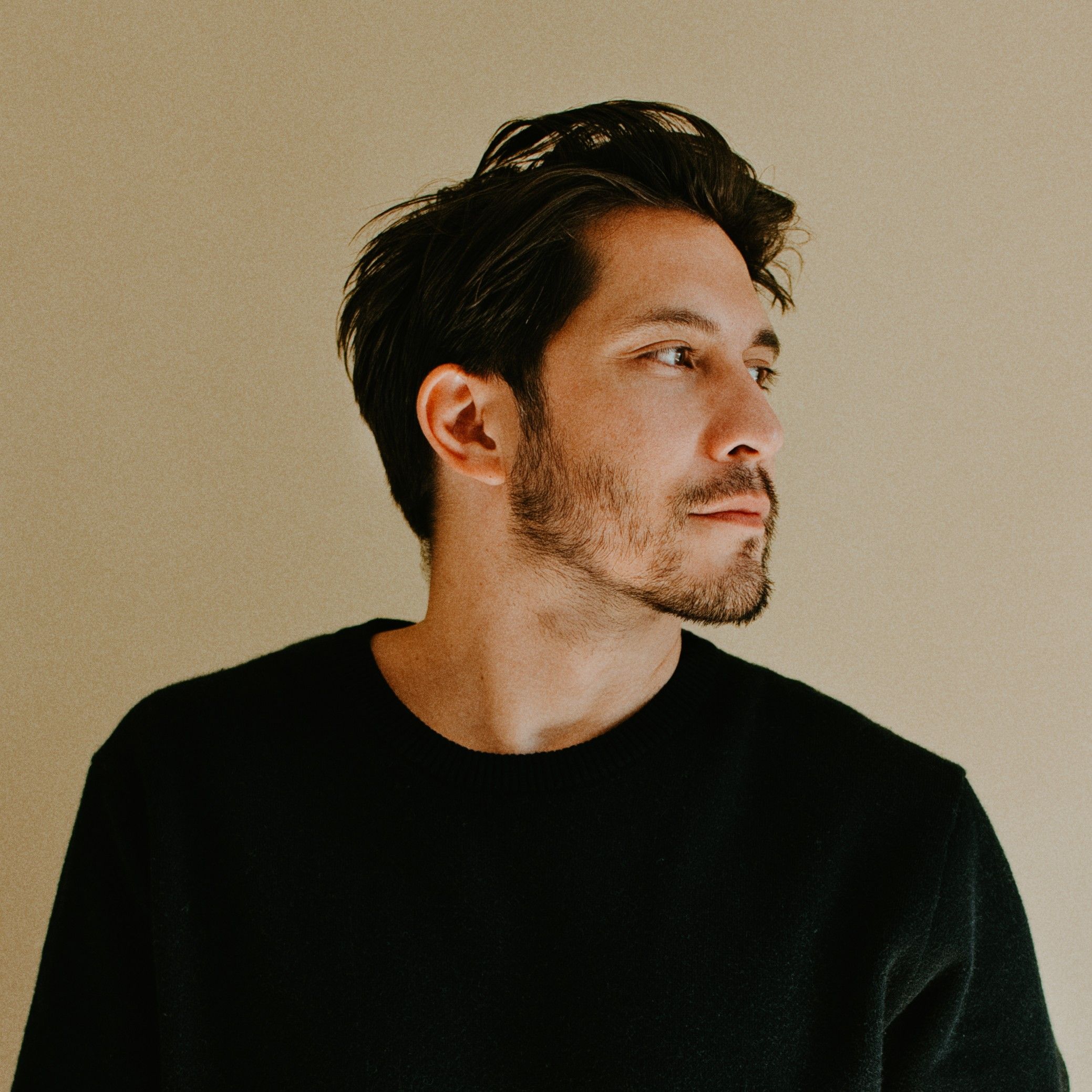 Young man in black t-shirt standing against beige wall, looking sideways in a minimalist portrait photo.