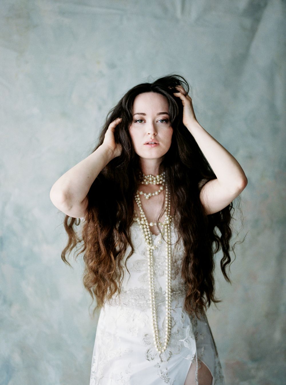 Ethereal portrait features long wavy brown hair, white CLAIRE PETTIBONE dress and layered pearl necklaces against textured gray backdrop.