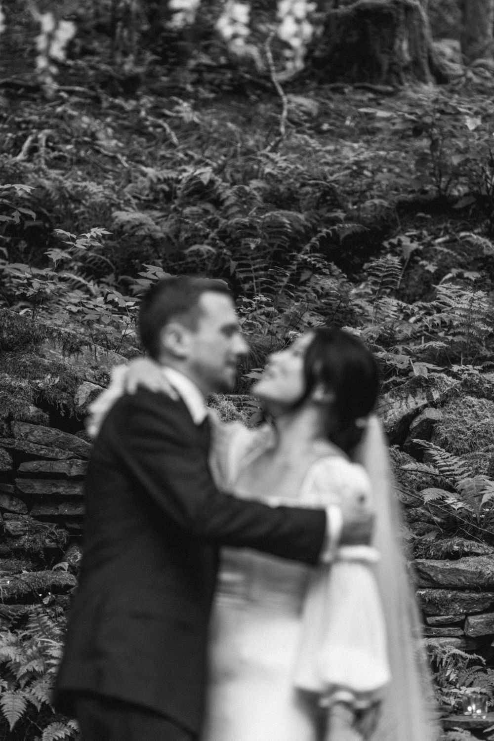 A romantic black and white wedding photo of a couple embracing in front of ferns and forest foliage.