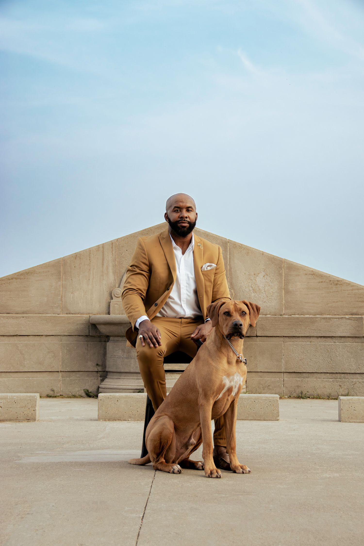 Bald man in tan suit sitting on stone steps with Rhodesian Ridgeback dog under blue sky.