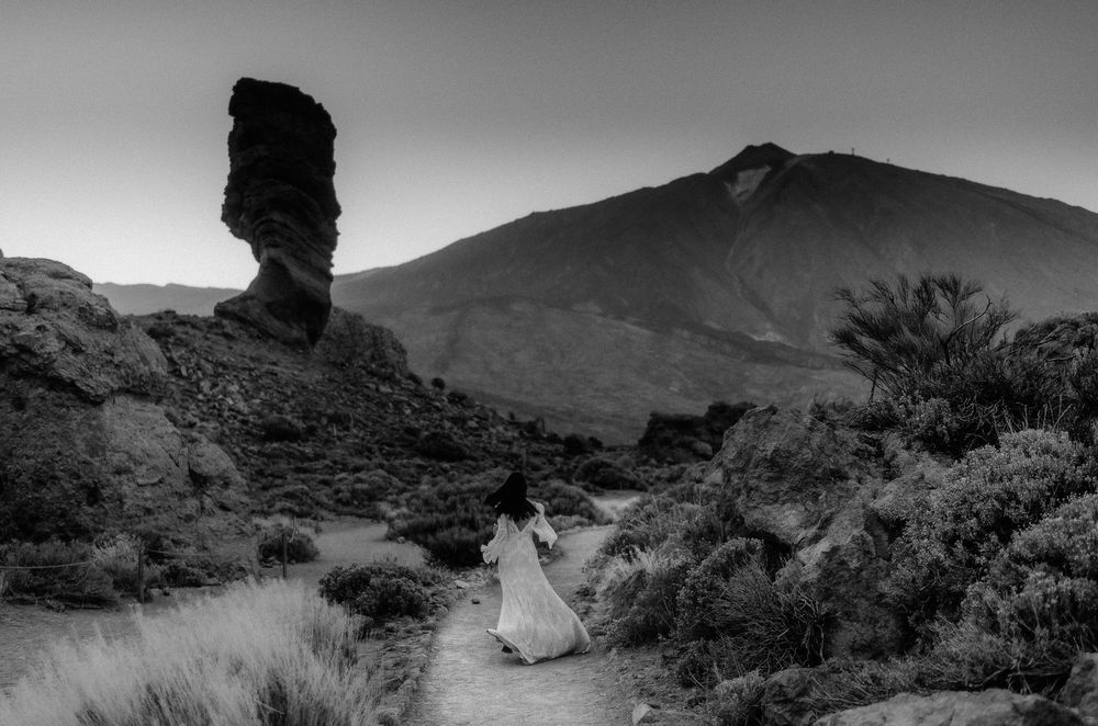 Black and white photo of a woman in white dress on a rocky path near Roque Cinchado with Mount Teide in background.