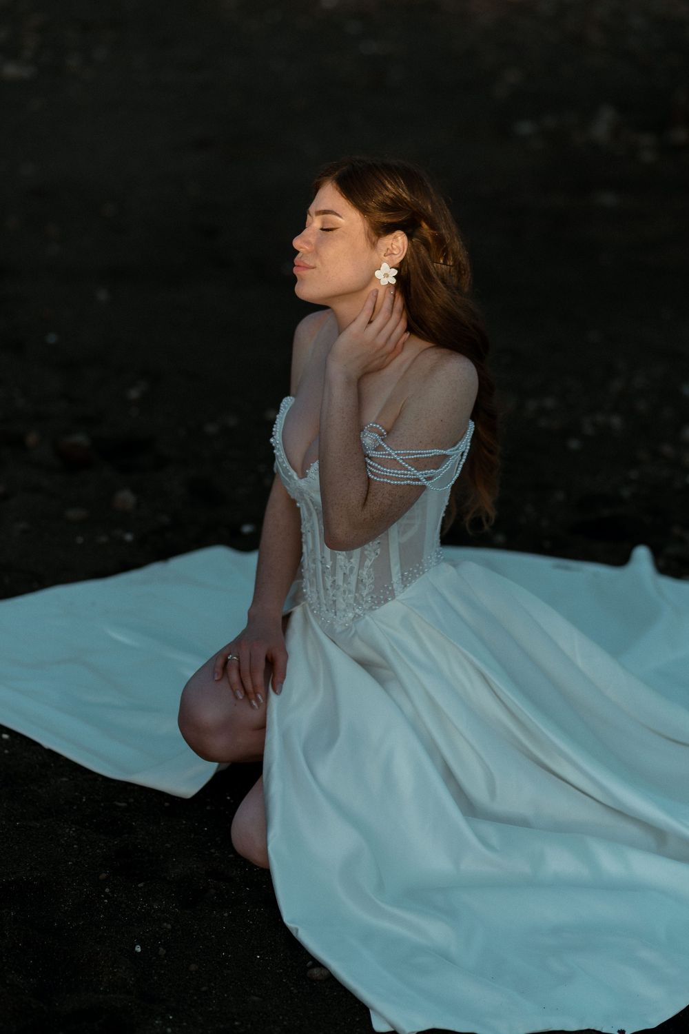 Woman in white off-shoulder wedding gown sitting gracefully on dark beach, eyes closed, wearing floral earrings.