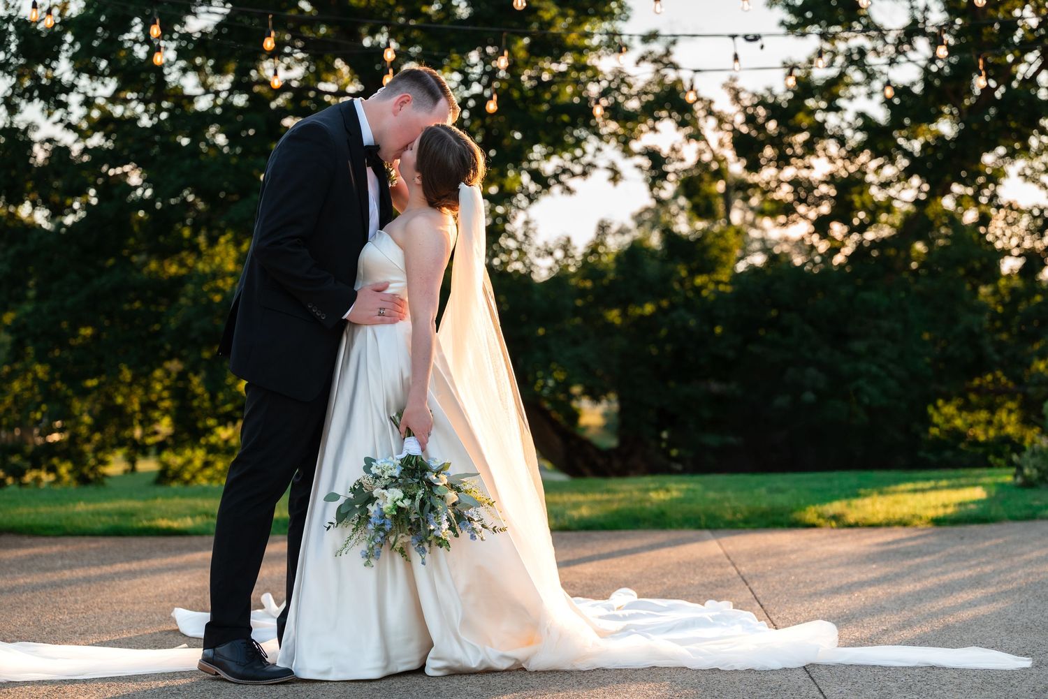 Bride and groom share a romantic kiss outdoors at golden hour, with lush green trees and string lights in the background.