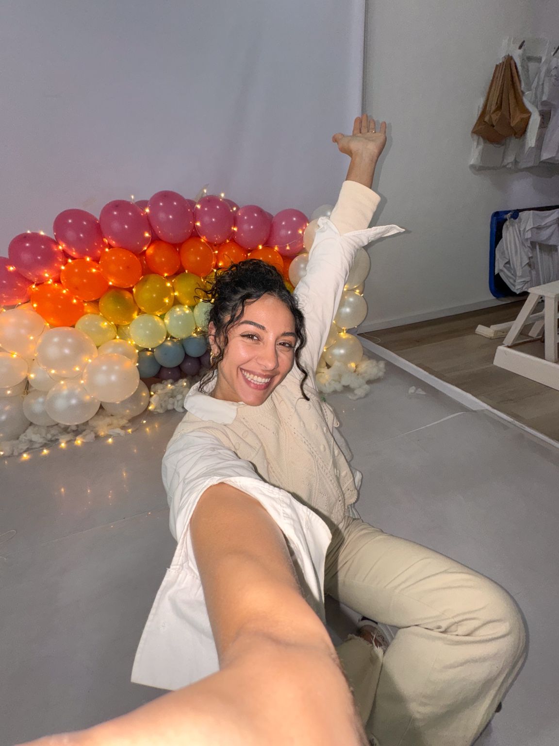 Woman smiling and posing in front of a colorful rainbow balloon wall with fairy lights on the floor.