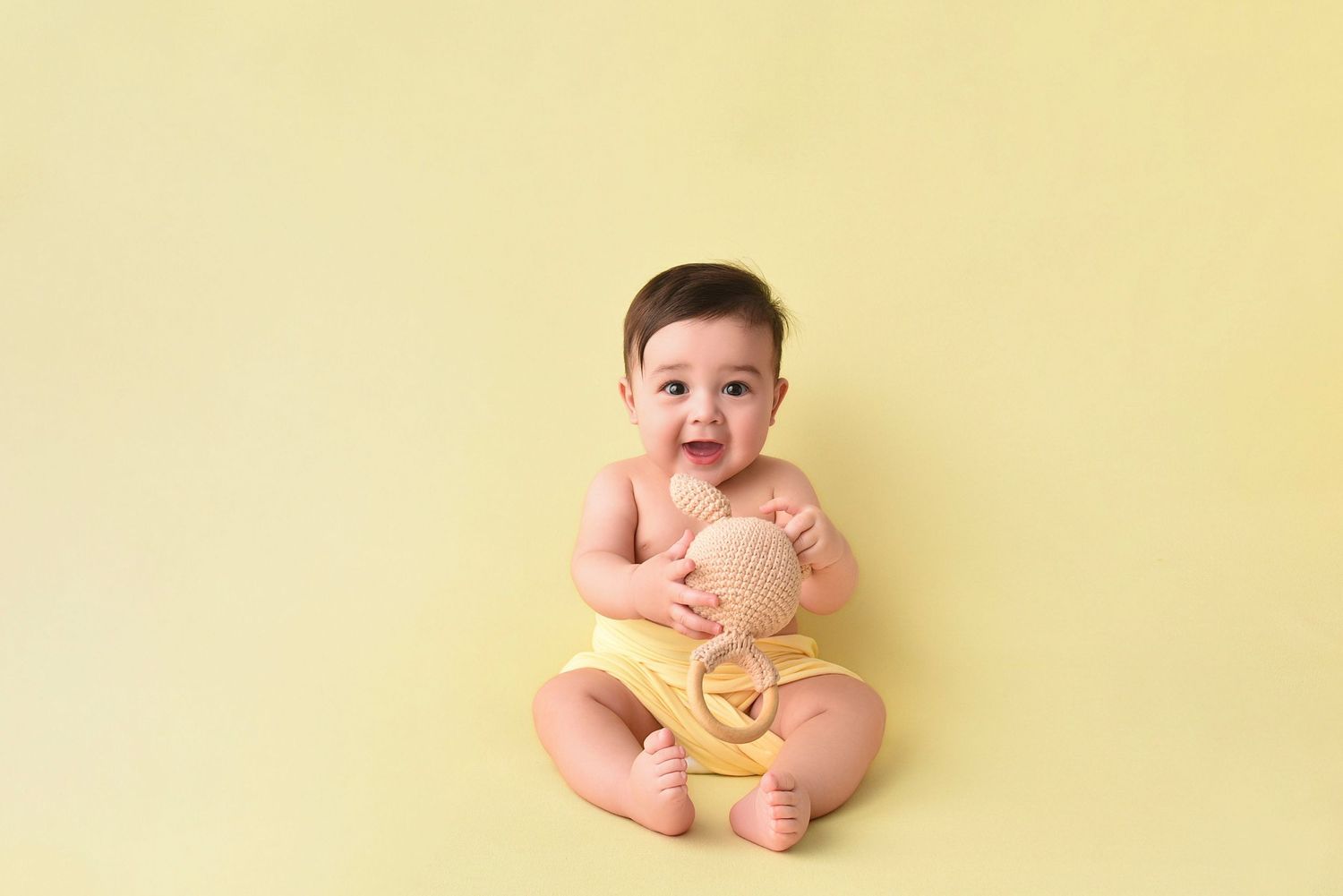 Smiling baby in yellow romper sitting on yellow background holding a wooden toy.