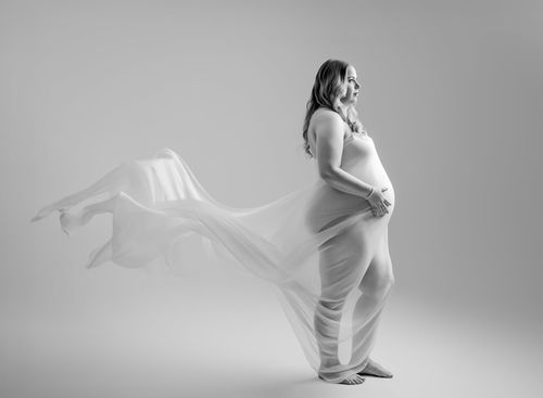 Black and white maternity photo of pregnant woman draped in flowing white fabric against light background.