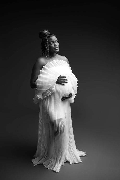 Black and white maternity photo of a pregnant woman in a white pleated gown cradling her baby bump in a studio.