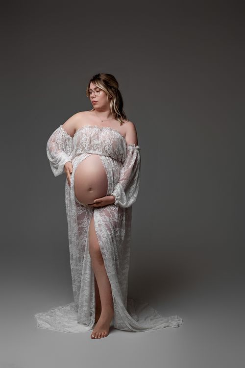 Pregnant woman in sheer white lace off-shoulder gown cradling baby bump in studio.
