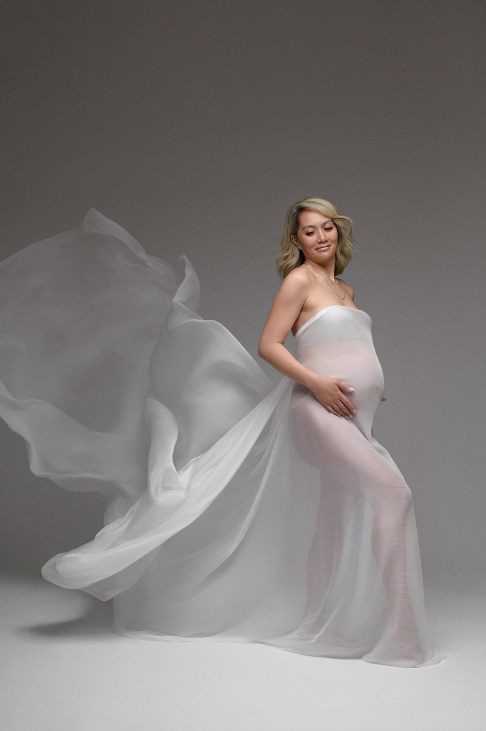 Pregnant woman wrapped in flowing white sheer fabric, posing gracefully in a studio maternity photography session.