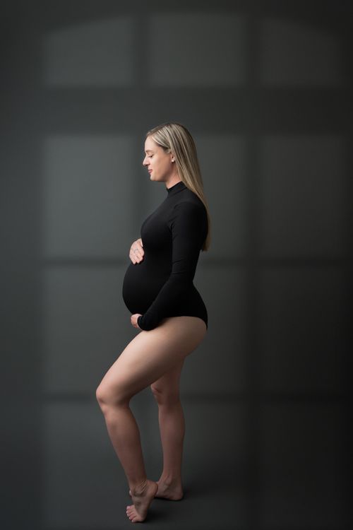 Pregnant woman in black long-sleeve bodysuit cradling her baby bump, standing sideways in a studio.