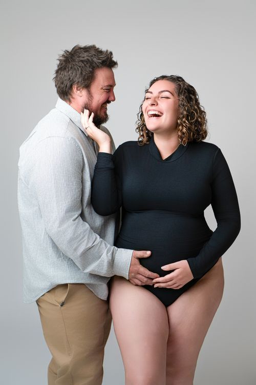 Happy pregnant couple laughing together in studio, woman in black bodysuit cradling baby bump with partner beside her.