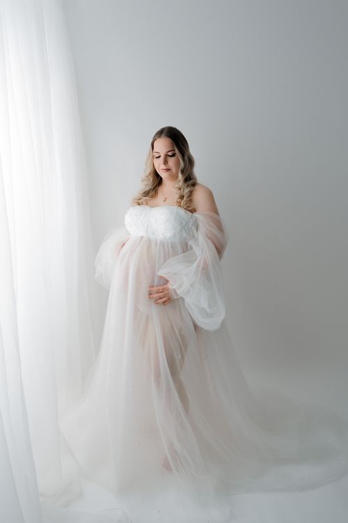 Pregnant woman in white floral bodice and sheer blush tulle gown poses near bright white curtains.