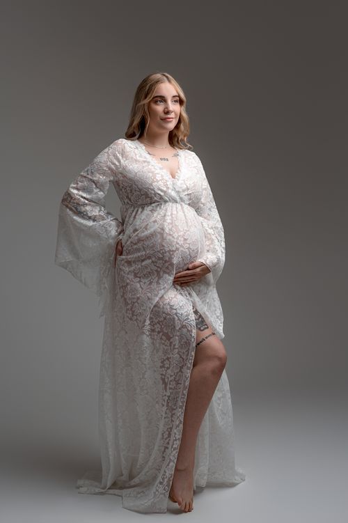 Smiling pregnant woman in sheer white lace maxi robe cradles baby bump in soft studio lighting.