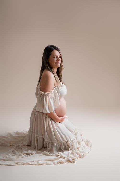 Pregnant woman in off-shoulder cream dress kneels serenely, cradling her baby bump in a soft-lit maternity portrait.