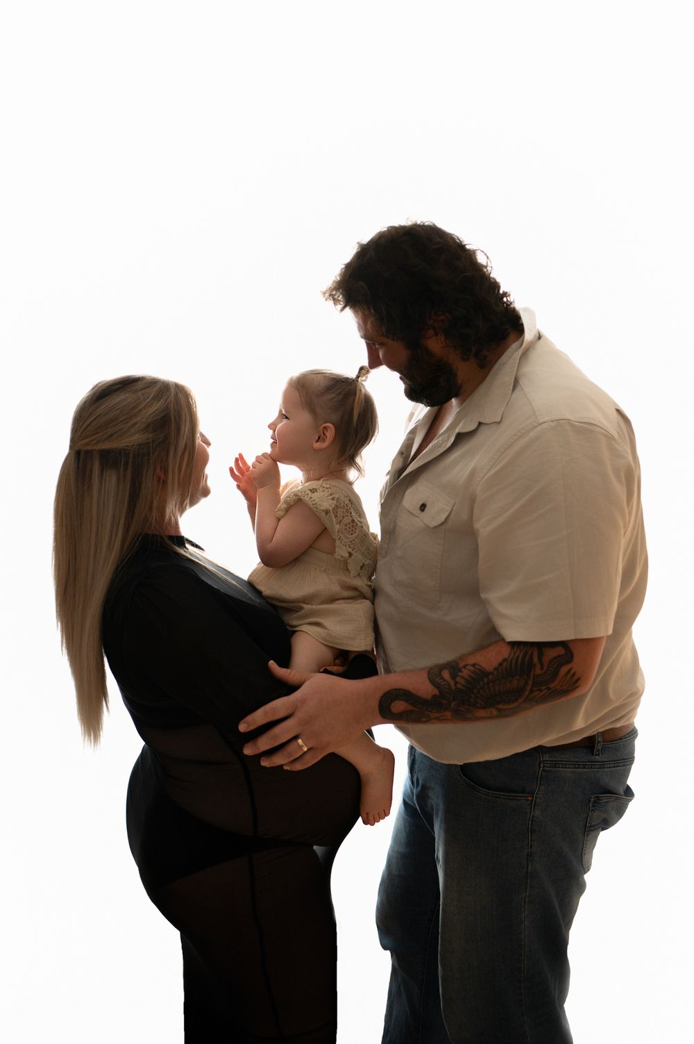 Pregnant woman in black dress with tattooed husband holding toddler daughter in a warm family maternity portrait.