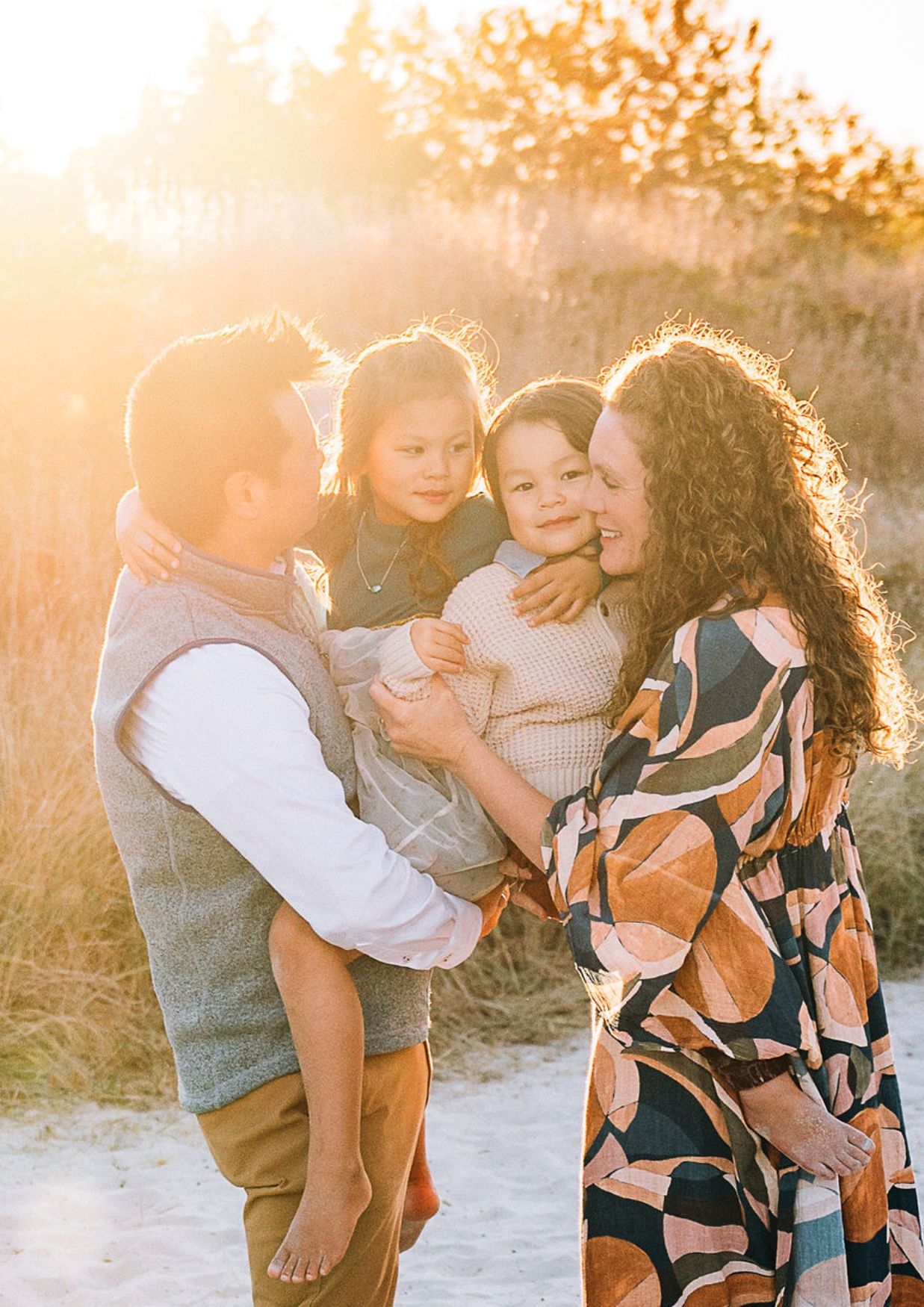 Family of four sharing a warm embrace on a sunlit beach, with golden hour light glowing behind them.