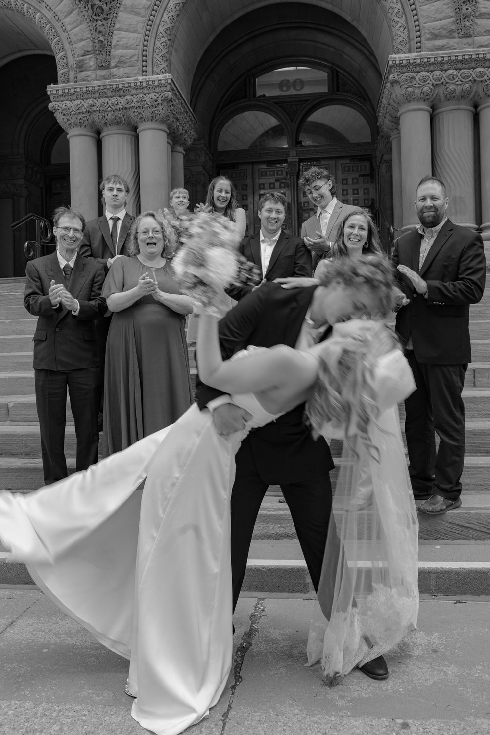 Groom dips bride dramatically on courthouse steps as wedding guests cheer in black and white photo.