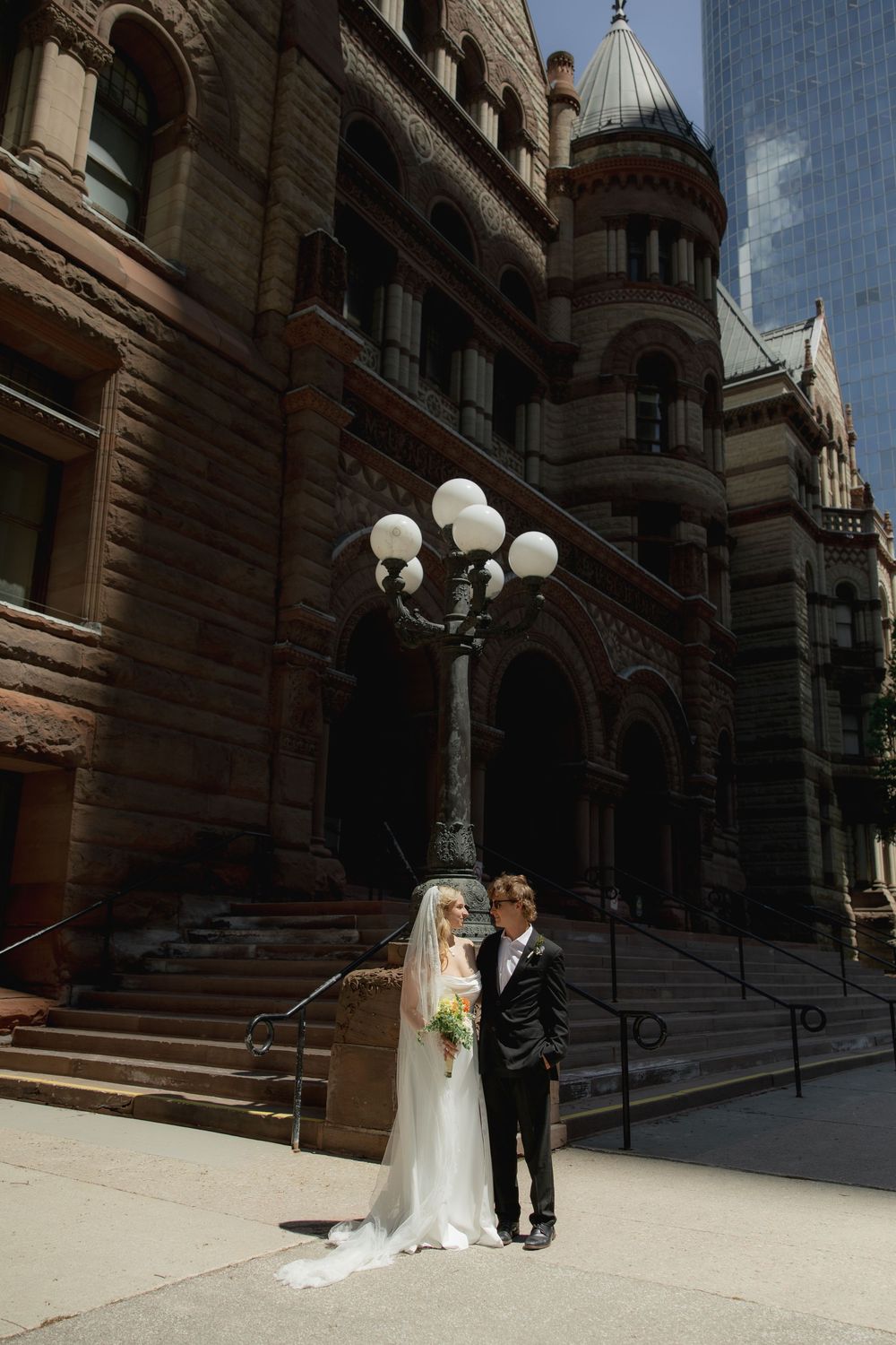 Bride and groom standing together near an ornate vintage street lamp outside a historic stone building.